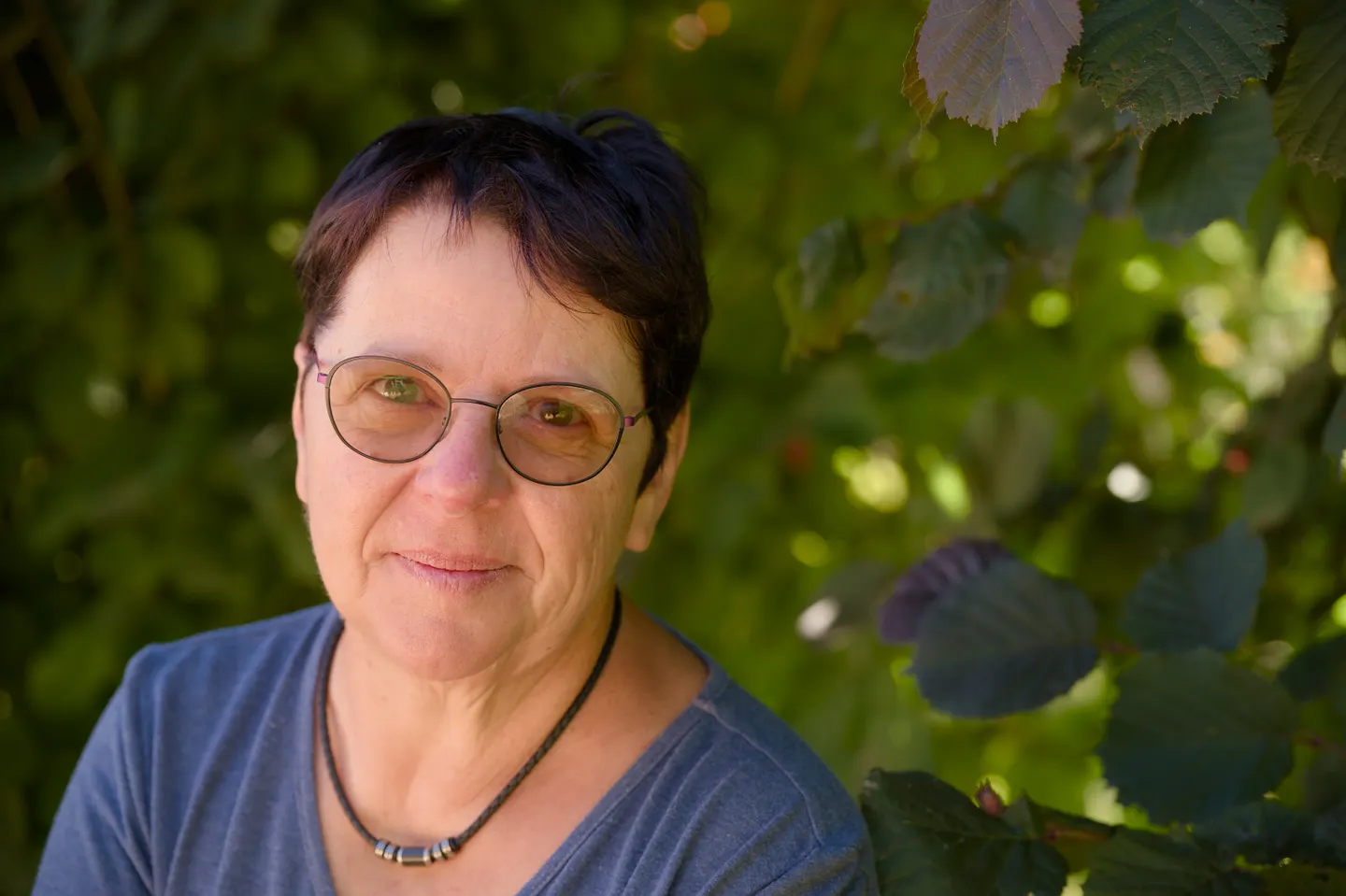 Retrato provisional de una mujer con gafas y camiseta azul ante un fondo de hojas verdes.