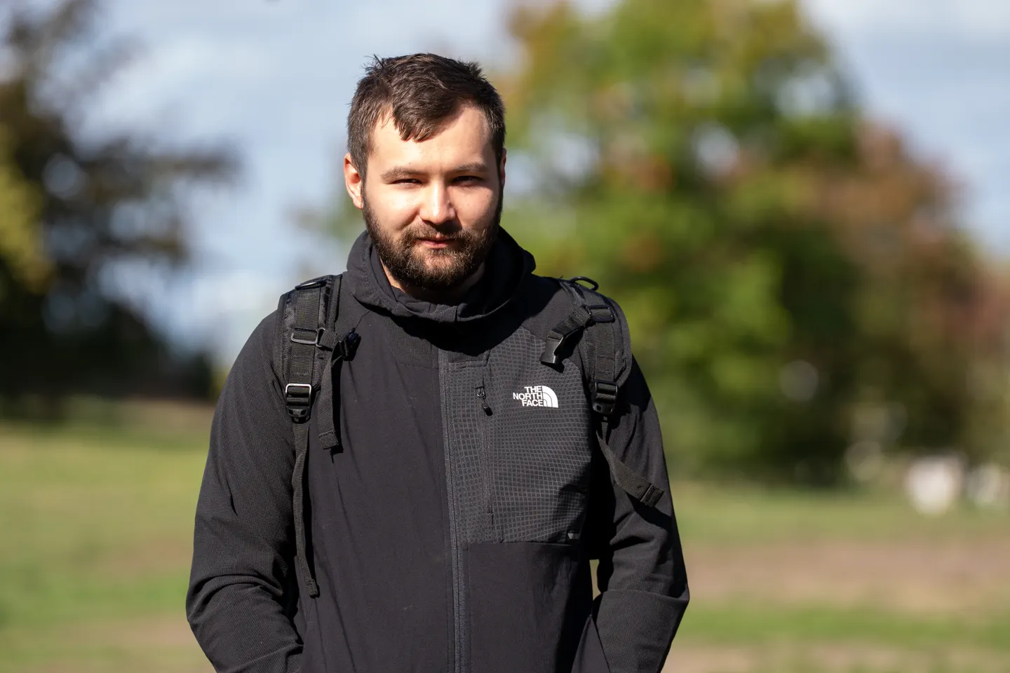 Provisional portrait of a man wearing a black jacket and backpack in a park.
